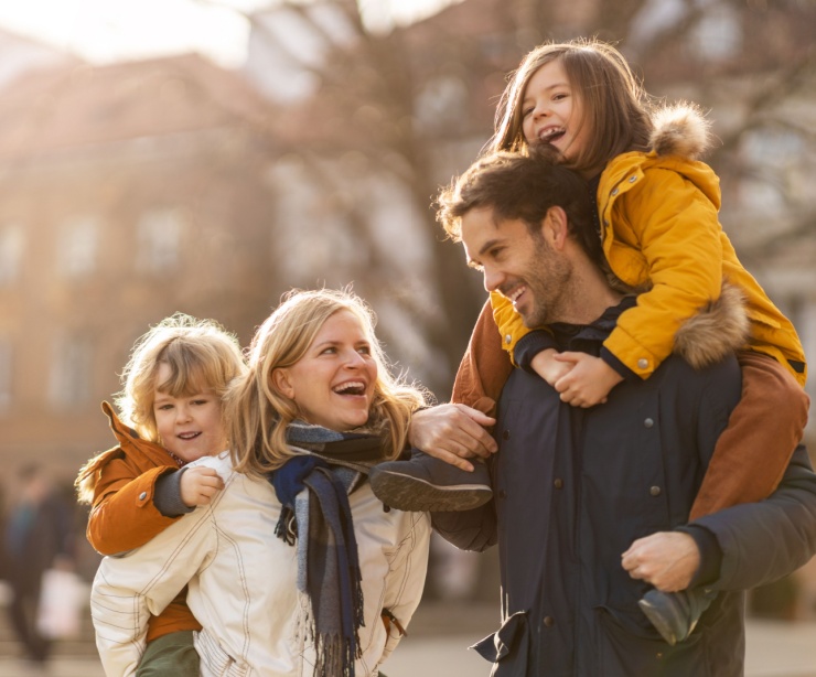 A husband and wife both give piggybacks to their young children in the middle of a city.